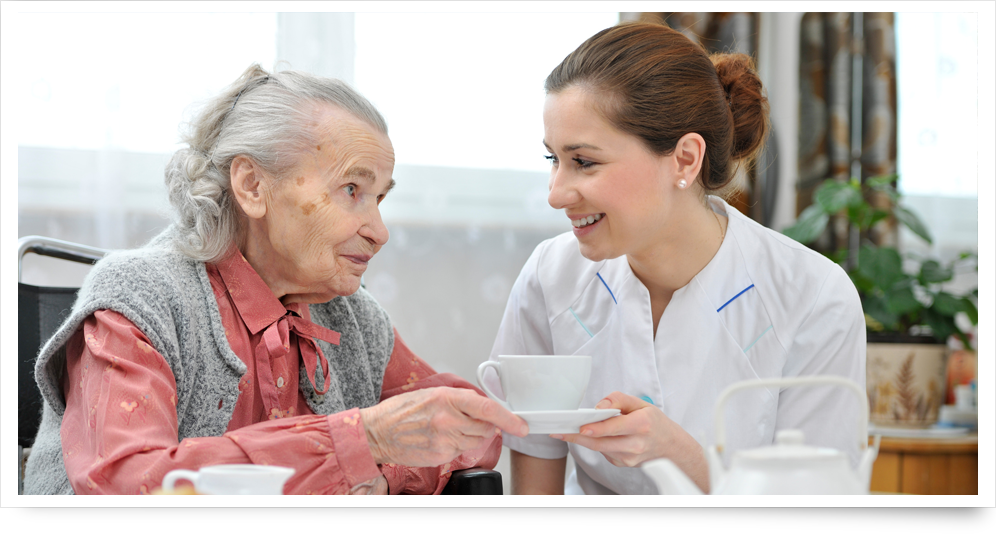 Junge Pflegerin reicht einer Seniorin eine Tasse Tee